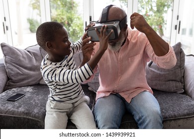 Front view of African american Grandson helping his grandfather to wear virtual reality headset in living room at home