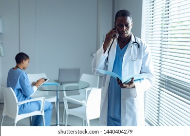 Front View Of African American Male Doctor Talking On Mobile Phone While Mixed Race Doctor Working On Desk In Hospital