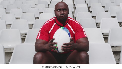 Front view of African American male rugby player sitting with rugby ball in stadium. He is looking away 4k - Powered by Shutterstock