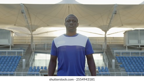 Front view of an African American male field hockey player, wearing a blue team strip, walking towards the camera, holding hockey stick on a hockey pitch looking at camera, on a sunny day - Powered by Shutterstock