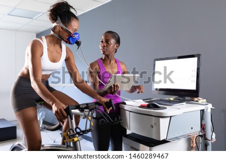 Front view of an African-American athletic woman doing a fitness test using a mask connected to a monitor while riding an exercise bike and an African-American woman monitoring her inside a room at a