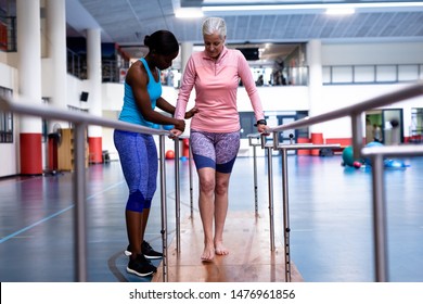 Front view of African-american female physiotherapist helping disabled Caucasian senior woman walk with parallel bars in sports center. Sports Rehab Centre with physiotherapists and patients working - Powered by Shutterstock