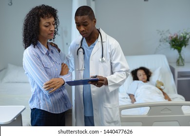 Front View Of African-American Male Doctor Discussing Over Clipboard With Mixed Race Woman In The Ward At Hospital. In The Background Mixed Race Child Sleeping