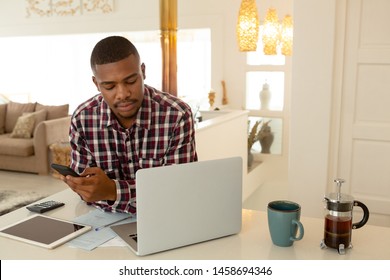 Front View Of African-american Man Using Mobile Phone And Laptop At Table In A Comfortable Home. Working From Home In Quarantine Lockdown. Social Distancing Self Isolation