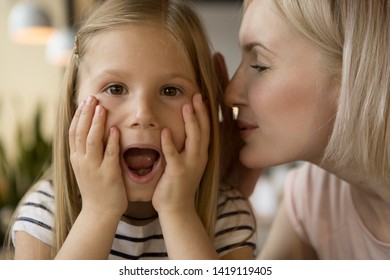 Front view of amazed little girl with open moth looking at camera while pretty mother whispering secret. Shocked daughter keeping hands on face. Concept of surprise and gossiping.