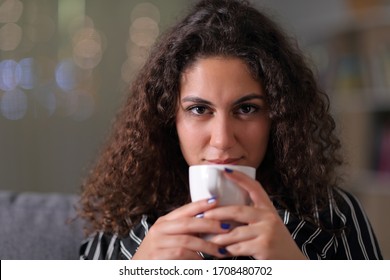 Front view of an arab woman with coffee cup looking at camera sitting on a couch at night at home