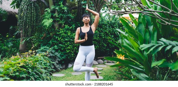 Front View Of Asian Girl Practicing Yoga In Tree Pose Outdoor. Harmony And Mental Health. Young Athletic Woman Wearing Sportswear And Barefoot Looking At Camera In Tropical Garden. Bali Island