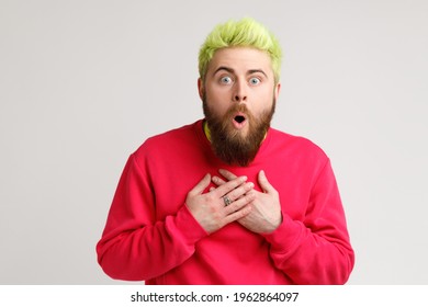Front view of astonished man in casual sweater standing with mouth open in surprise, has shocked expression, hears unbelievable news, hands on chest. Indoor studio shot isolated on gray background.