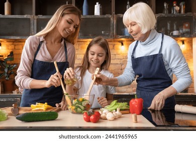 Front view of beautiful little girl that pours oil in salad while young mother and grandmother helping her to mix it. Three generations of women cooking healthy food at home - Powered by Shutterstock