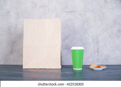 Front view of blank take away food bag, cookies on saucer and green coffee cup on wooden surface and concrete background. Mock up - Powered by Shutterstock