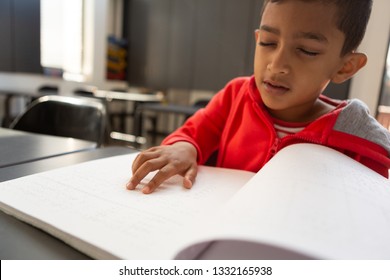 Front view of blind mixed-race schoolboy reading a braille book at desk in a classroom at elementary school