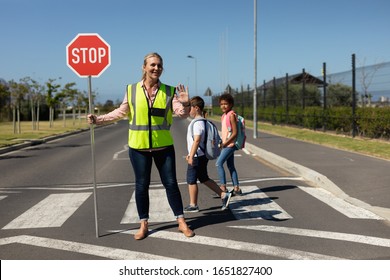 Front View Of A Blonde Caucasian Woman Wearing A High Visibility Vest And Holding A Stop Sign, Standing In The Road And Stopping Traffic On A Pedestrian Crossing, While Two Schoolchildren Cross The