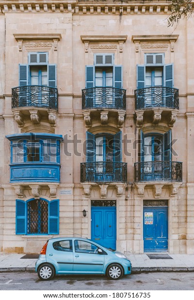 Front view\
of blue car in front of traditional Maltese house with blue covered\
balconies in historical part of the old town of Valletta in Malta.\
Vintage picturesque tourist postcard.\

