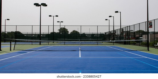 Front view of blue and green tennis courts with lights surrounded by tall black fencing. - Powered by Shutterstock