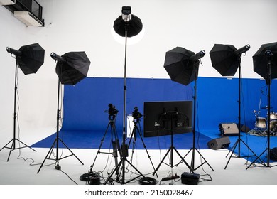 Front View Of Blue Screen Backdrop With A White Pedestal Was Set For Filming, With The Back Of Many Light Equipments, Video Camera On Tripods, And Monitor, Setup In Front Of Blue Screen In The Studio.