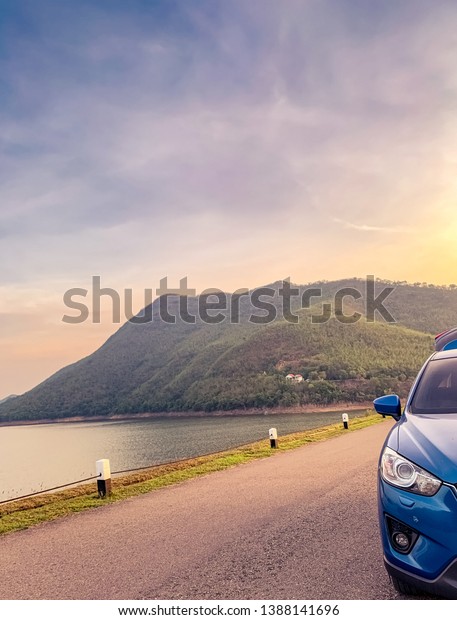 Front view of blue SUV car with opened car truck\
parked on asphalt road at dam. Summer road trip travel. Car parked\
on the road in front of the mountain beside reservoir. Summer\
vacation with nature.