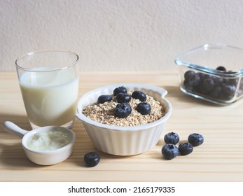 Front View Of Breakfast With Oat Or Granola In White Bowl, Fresh Blueberries,  Glass Of Milk And Yogurt  On Wooden Table. Healthy Breakfast Concept.