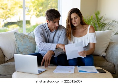 Front View Of A Caucasian Couple At Home, Sitting In The Living Room On A Couch Looking At Paperwork And Using A Laptop Computer, The Woman Holding A Document And The Man Pointing