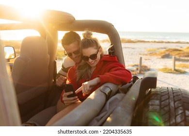 Front view of a Caucasian couple inside an open top car, using their smartphone and smiling. Weekend beach vacation, lifestyle and leisure. - Powered by Shutterstock