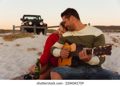 Front view of a Caucasian couple reclining on the beach, with an open top car in the background, the man holding the guitar, the woman hugging her partner and holding a bottle of beer - Powered by Shutterstock