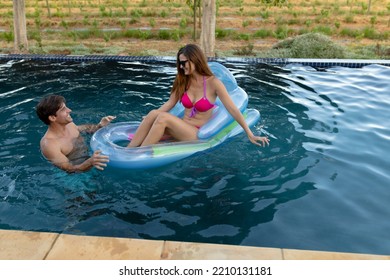 Front view of a Caucasian couple in a swimming pool, the man standing and holding onto an inflatable pool lounger and the woman sitting on it wearing sunglasses and smiling - Powered by Shutterstock