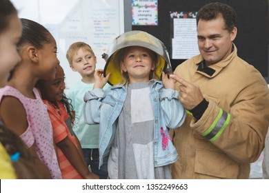 Front View Of A Caucasian Firefighter Teaching To School Kids About Fire Security While One Of Them Has A Fire Helmet Over Her Head