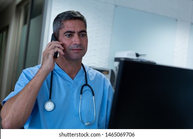 Front view of Caucasian male surgeon talking on mobile phone while working on computer at desk in hospital. Healthcare workers in the Coronavirus Covid19 pandemic - Powered by Shutterstock