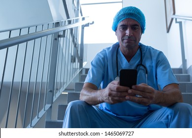 Front view of Caucasian male surgeon using mobile phone while sitting on stairs at hospital. Healthcare workers in the Coronavirus Covid19 pandemic - Powered by Shutterstock