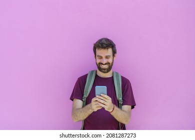 Front View Of Caucasian Man Holding A Phone On Pink Background. Horizontal View Of Latin American Man Using Technology Isolated On Pink Wall. People And Travel Concept.