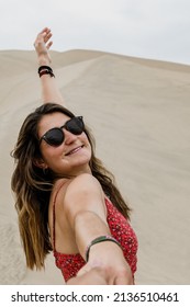 Front View Of A Caucasian Woman With Sunglasses Standing Up In The Dune Of A White Desert Looking At The Camera, Smiling And Raising One Arm, Held By A Man Hand. Vertical. With Copy Space.