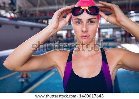 Front view of a Caucasian woman wearing a swimsuit and a pink swimming cap holding goggles  while standing by an olympic sized pool inside a stadium
