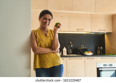 Front view of a cheerful woman leaning her shoulder against the wall in the kitchen - Shutterstock ID 1815220487
