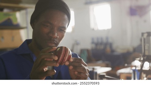 Front view close up of an African American man working in the workshop at a factory making cricket balls, holding and carefully inspecting a finished cricket ball in the sunlight, slow motion - Powered by Shutterstock