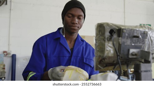 Front view close up of an African American man wearing gloves, overalls and a hat working at a factory making cricket balls, preparing a mixture for moulding into the core of the balls - Powered by Shutterstock