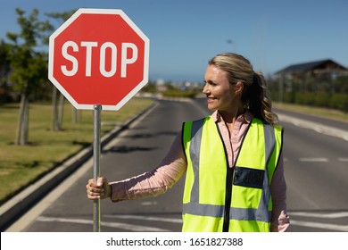 Front View Close Up Of A Blonde Caucasian Woman Wearing A High Visibility Vest And Holding A Stop Sign, Standing In The Road And Looking To The Side On A Pedestrian Crossing, Waiting To Help Children