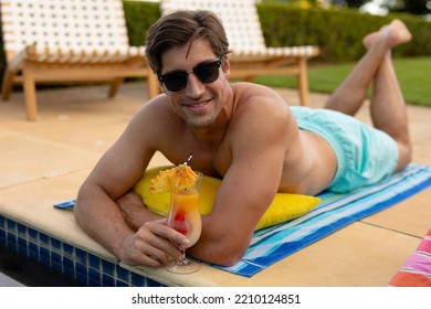 Front View Close Up Of A Caucasian Man Wearing Beachwear And Sunglasses In A Garden, Lying Beside A Swimming Pool, Holding A Cocktail, Sunbathing, Looking To Camera And Smiling