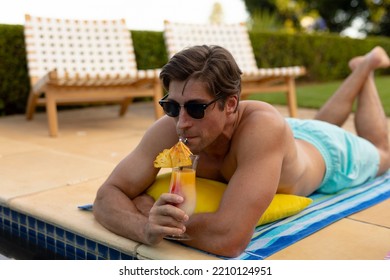 Front View Close Up Of A Caucasian Man Wearing Beachwear And Sunglasses In A Garden, Lying Beside A Swimming Pool, Sunbathing And Drinking A Cocktail 