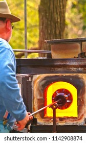 Front View, Close Distance Of A Senior, Male Artisan Rolling Glass In The Hot Fire Of An Out Door Furnace And Preparing To Blow The Glass Into A Vase Or Glass Object At A Summer Fair In Pennsylvania 