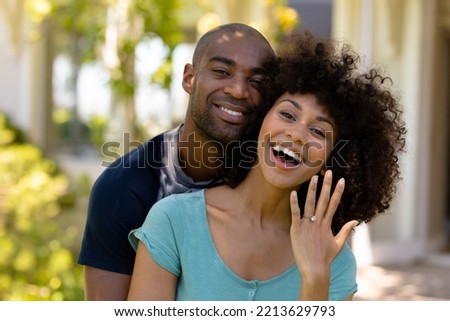 Front view close up of a mixed race couple standing in the garden, embracing and smiling to camera, the woman showing the engagement ring that the man has just given her