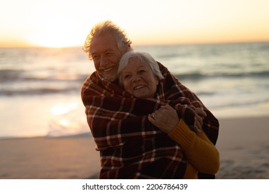Front view close up of a senior Cacuasian couple standing on the beach at sunset with a blanket over their shoulders, smiling to camera and embracing, with the sea in the background - Powered by Shutterstock