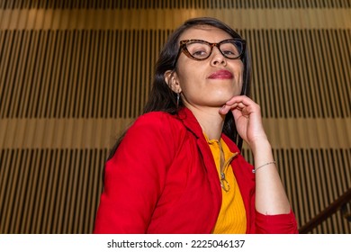 Front view, close-up portrait of an attractive young Hispanic woman, wearing glasses, red jacket, jeans with one hand under her chin against a background of wooden poles - Shutterstock ID 2225046137