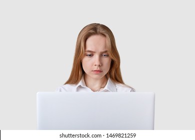 Front View Of Concentrated Adult Girl Sitting In Front Of White Portable Laptop, Having Online Work, Looks At Computer Screen, Wearing Blouse, Has Straight Fair Hair, Using Wireless Internet.