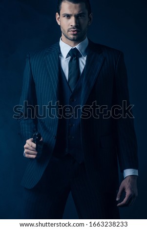 Front view of confident gangster aiming weapon and looking at camera on dark blue background