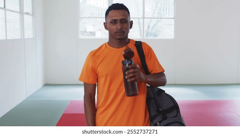 Front view of a confident happy teenage biracial male judoka wearing orange t-shirt, standing in the gym with gym bag, drinking water from plastic bottle after a judo training - Powered by Shutterstock