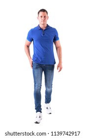 Front View Of Confident Young Adult Man Walking Forwards And Looking At Camera In Blue Polo Shirt. Full Body Isolated On White Background.