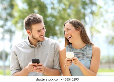 Front view of a couple or friends joking and using smart phones on line outdoors in a park