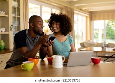 Front view of a  couple at home sitting in the kitchen, smiling at each other and using mobile phone and a laptop  - Shutterstock ID 2213633755
