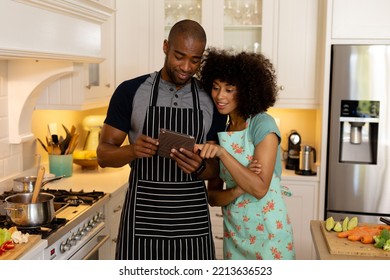 Front view of a couple at home, standing in the kitchen wearing aprons, smiling and using a tablet computer together - Powered by Shutterstock