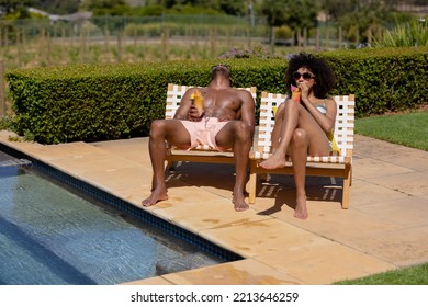 Front view of a couple sitting in the garden by a swimming pool wearing beachwear, sunbathing on sunloungers, the woman in sunglasses, drinking a cocktail - Powered by Shutterstock