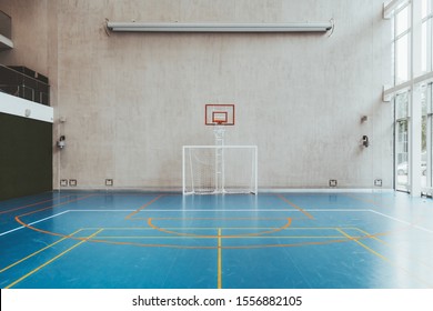 Front view of the court in the gymnasium hall; an indoor modern office stadium with a basketball basket and hoop, football goal, blue floor, a concrete wall with an undeployed projection screen above
